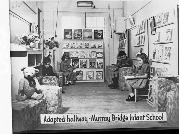 Eine Gruppe von Kindern sitzt auf Stühlen in einem Raum mit Bücherregalen, ein Teddybär links, eine Vase auf dem Tisch und Rahmen an der Wand. Unten steht der Text "Angepasster Flur Murray Bridge Infant School".