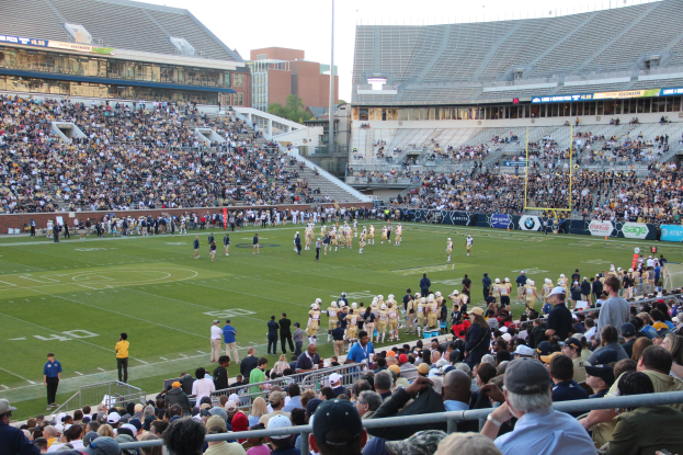 Fußballspiel im Gange mit Spielern auf dem Feld und Zuschauern in den Stadionrängen unter einem klaren Himmel.