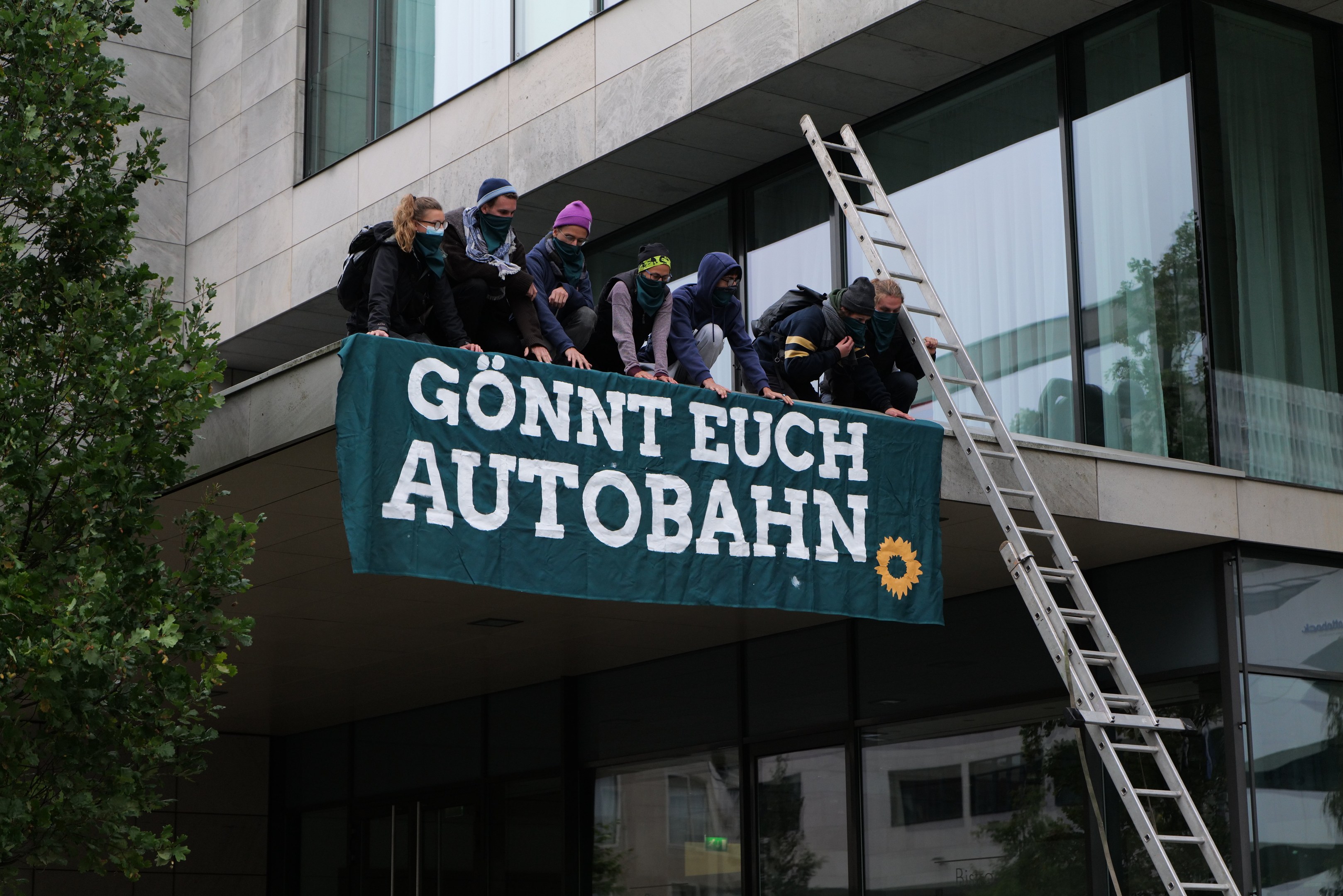 Eine Gruppe von Menschen auf einem Gebäude, die eine Fahne halten, mit einem Baum links und Glasfenstern im Hintergrund, die scheinbar für die deutsche Regierung protestieren.