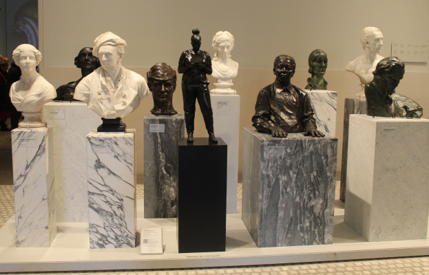 Eine Gruppe detaillierter Büsten-Skulpturen in einem Museum ausgestellt, mit Besuchern im Hintergrund und einer Wand dahinter.