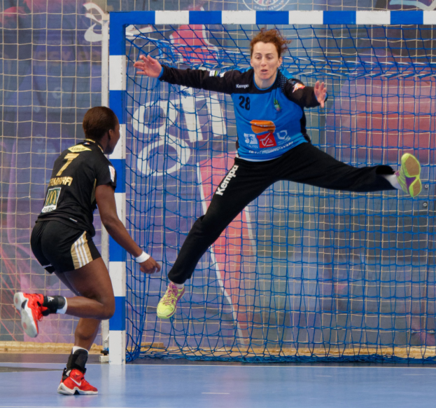 Zwei Frauen beim Handballspiel auf einem Platz mit Tor und Banner im Hintergrund, beide tragen Sportschuhe.