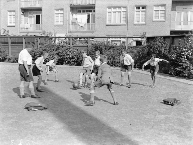 Schwarzes Foto von jungen Jungs, die Fussball auf einem grünen Feld mit einem Ball in der Mitte spielen, mit Gebäuden mit Fenstern und Balkonen im Hintergrund und Text unten.