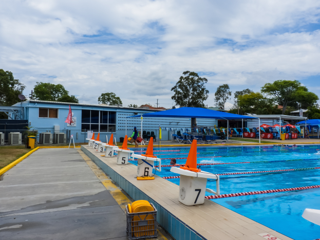 Ein großes Schwimmbad mit schwimmenden Menschen, Bahnmarkierungen, Verkehrskegeln, Stühlen, Sonnenschirmen, einem Gebäude mit Fenstern, einer Fahne, Bäumen und einem bewölkten Himmel im Hintergrund.