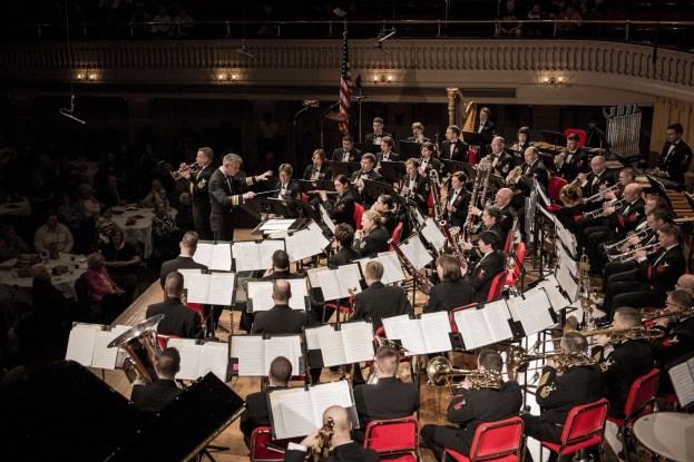 Große Militärmusikformation in einem Konzertsaal mit sitzenden und stehenden Musikern, Notenpulten und einer Flagge an der Rückwand.