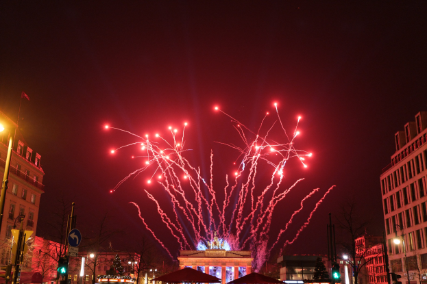 Eine Straße in einer Stadt mit Gebäuden, Bäumen, Laternen, Ampeln, Schildern, Zelten und Menschen, mit Feuerwerk, das den Himmel erhellt, während einer Silvesterfeier in Berlin.