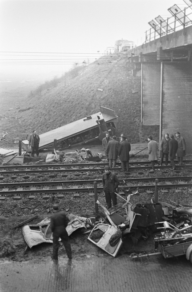 Eine Schwarz-Weiß-Szene eines Zugunglücks auf den Schienen, mit einer Gruppe von Menschen um das Wrack herumstehend, einem Fahrzeug im Vordergrund und einer Brücke, Polen, Drähte und Himmel im Hintergrund.