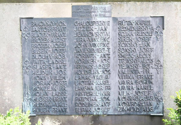 Ein Steinplatten auf einer Wand mit eingravierten Text, umgeben von Pflanzen unten, wahrscheinlich ein Denkmal für Holocaust-Opfer.