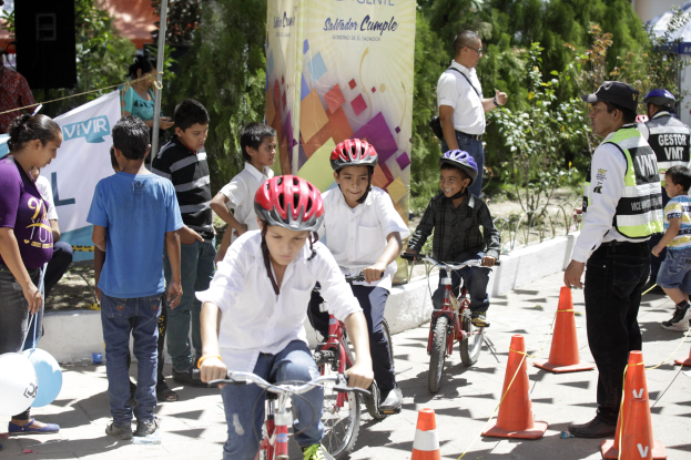 Kinder fahren mit Fahrrädern eine Straße entlang, vorbei an Verkehrskegeln, einige tragen Helme, andere stehen daneben, mit einer Fahne, Bäumen und Gebäuden im Hintergrund.