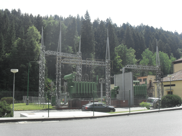 Eine elektrische Umspannstation am Straßenrand, umgeben von einem Zaun, Masten, Lampen, Pflanzen, Gras und Bäumen, mit einem vorbeifahrenden Auto und Gebäuden im Hintergrund bei einem klaren blauen Himmel.