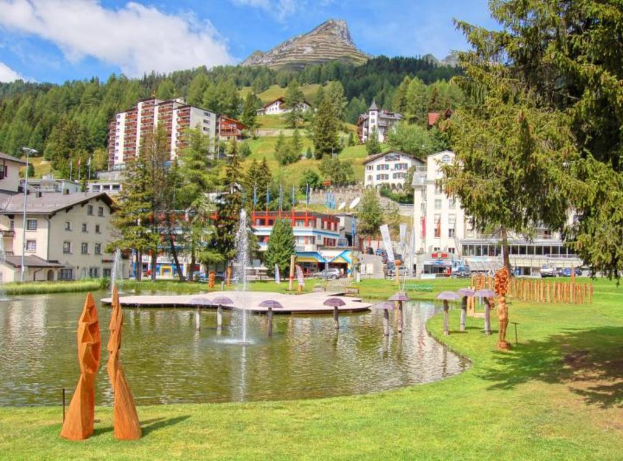 Ein zentraler Wasserplatz mit Fontänen, umgeben von Gebäuden, Bäumen und Bergen, mit Gras am Boden des Bildes.