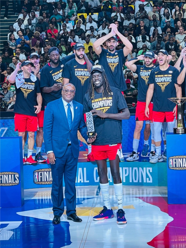 Eine Gruppe von Männern feiert auf einem Basketballfeld, zwei halten Trophäen, mit Zuschauern im Hintergrund und zwei Trophäen auf Podesten rechts.