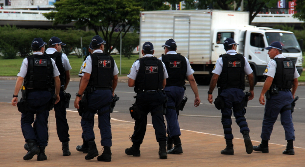 Eine Gruppe von Beamten in Uniform, einschließlich Mützen, Hemden, Schutzwesten, Uhren, Hosen und Schuhen, die auf einer Straße mit einem geparkten Lkw auf der rechten Seite neben einem Zaun, Gebäuden, Bäumen, Pflanzen, Pfählen und Schildern im Hintergrund und einer Brücke im oberen rechten Eck gehen.