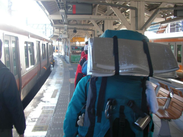 Ein Bahnhofsplatz mit Menschen neben einem Zug, einem Stand in der Mitte, einem weiteren Zug auf der rechten Seite und einer Bank in der unteren rechten Ecke.