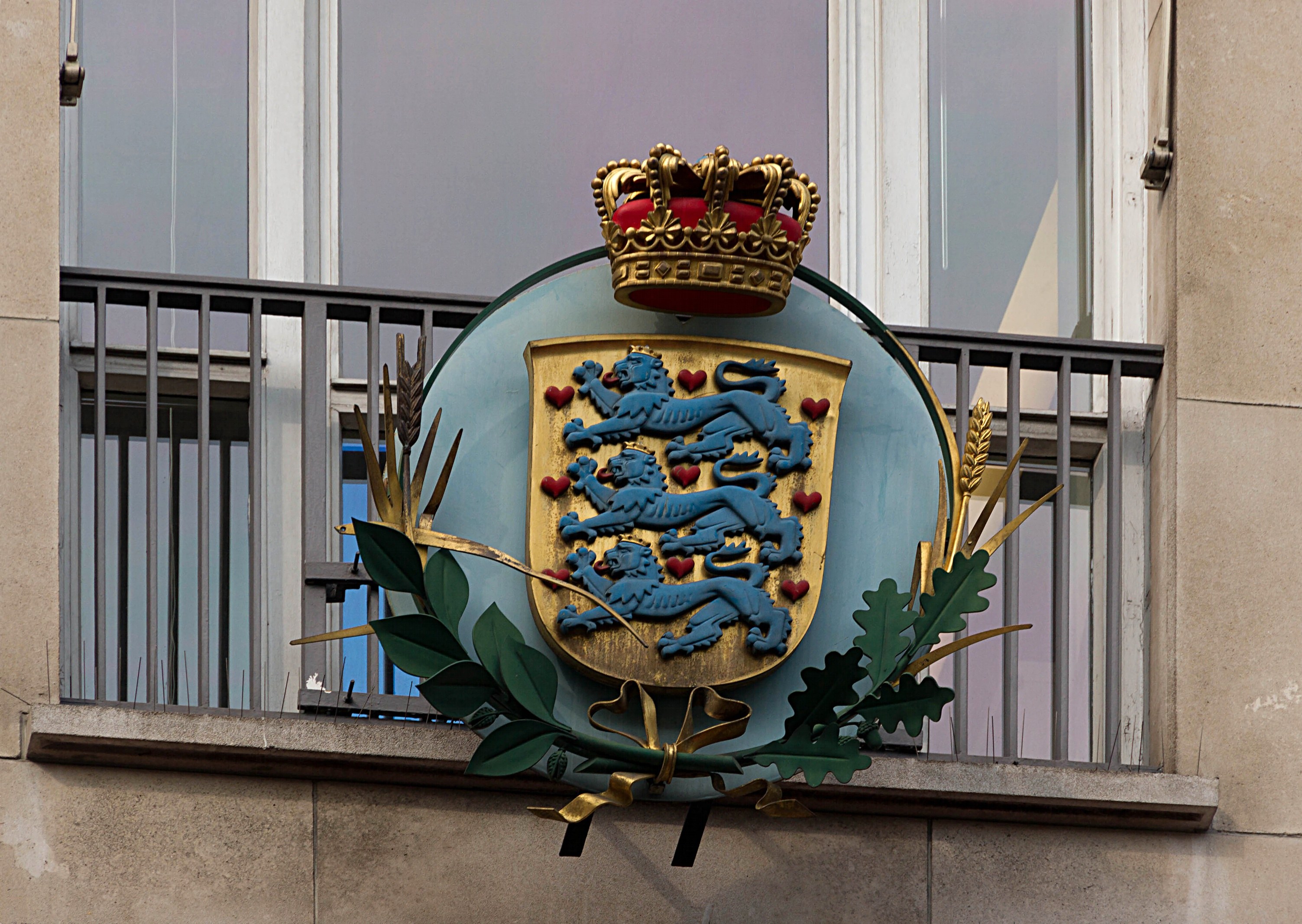 Das Wappen der britischen Königsfamilie, das auf der Außenseite eines Gebäudes dargestellt ist, begleitet von einem Logo mit Krone und Blättern, mit Fenstern und Metallgittern im Hintergrund.