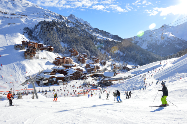 Eine Gruppe von Menschen, die eine schneebedeckte Piste in den französischen Alpen hinunterskifahren, umgeben von Bäumen, Häusern, Pfählen, Drähten und Bergen unter einem bewölkten Himmel mit strahlendem Sonnenschein.