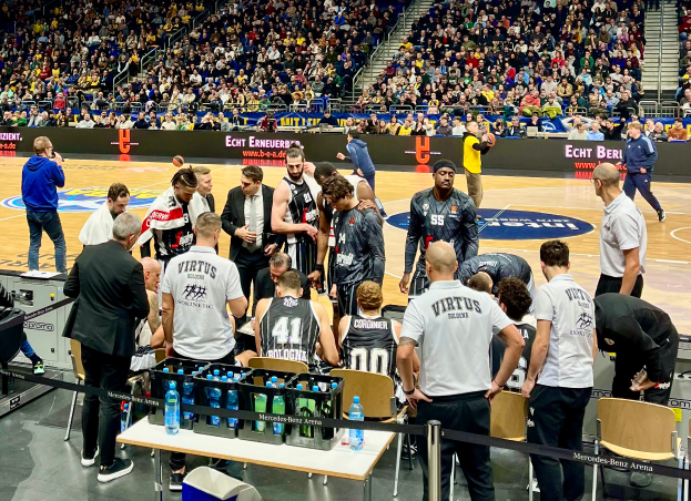 Eine Gruppe von Männern steht auf einem Basketballfeld umgeben von Zuschauern, mit einer Person, die links eine Kamera hält und Tischen mit Text im Hintergrund.