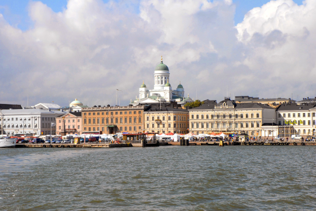 Ein braunes und weißes Gebäude mit vielen Fenstern steht vor einer Moschee mit einer grünen Kuppel, mit einem Hafen mit Booten und Fracht im Vordergrund und Meerwasser unten.
