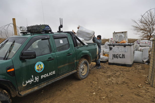 Polizei-Lkw auf einem Schotterweg mit sichtbarem Text und Logo neben einem Maschendrahtzaun geparkt, zwei Personen in der Nähe, eine hält eine Tasche, Hintergrund-Elemente wie Taschen, ein Baum, ein Pfahl und der Himmel.