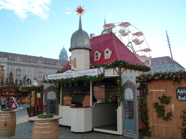 Kleines Gebäude mit rotem Dach und Riesenrad davor, umgeben von Festdekorationen und Menschen, mit Gebäuden, Bäumen und bewölktem Himmel im Hintergrund bei einem Oktoberfest-Event.