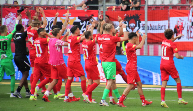 Gruppe von Fußballspielern, die ein Tor feiern, einige halten Flaschen und andere klatschen, mit Bannern, einem Zaun, Stühlen und einem Dach mit Lampen im Hintergrund.