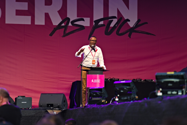 Ein Mann in einem weißen Hemd und braunen Hosen steht auf einer Bühne und hält ein Mikrofon, mit einem pinken Banner mit der Aufschrift "Berlin as Fuck" dahinter und einer Menge davor.