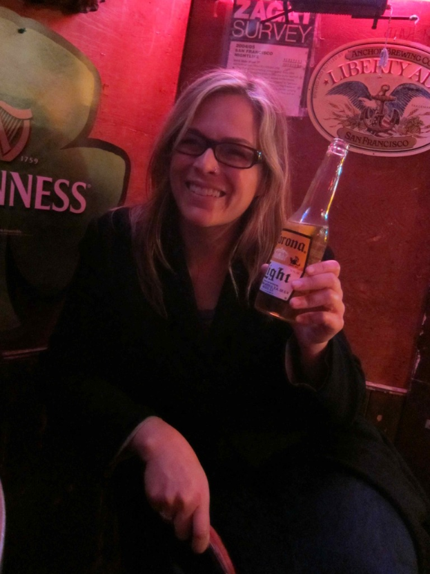 Eine Frau sitzt, lächelt, trägt eine Brille und einen Mantel und hält eine Bierflasche in ihrer linken Hand, vor einer Wand mit Plakaten und einem Logo im Hintergrund.