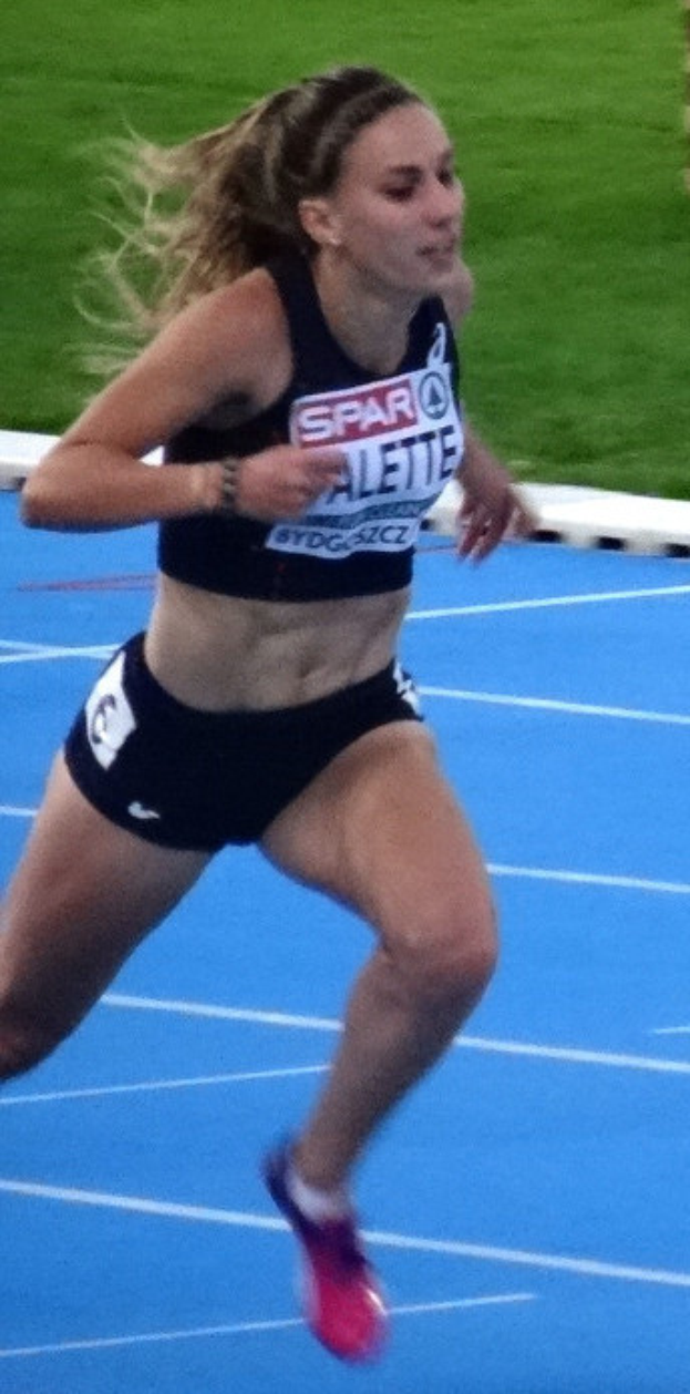 Eine Frau läuft auf einer blauen Laufbahn in einem Stadion, trägt ein Sport-BH-Oberteil und Shorts und ist von saftig grünem Gras umgeben.