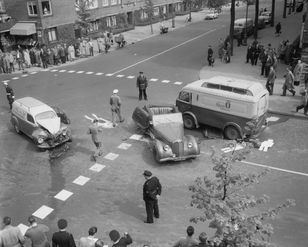 Schwarz-weiß-Szene eines Autounfalls an einer Straßenkreuzung mit mehreren Fahrzeugen, darunter ein Lieferwagen, und Zuschauern; Gebäude, Bäume, Strommasten und Schilder sind im Hintergrund zu sehen.