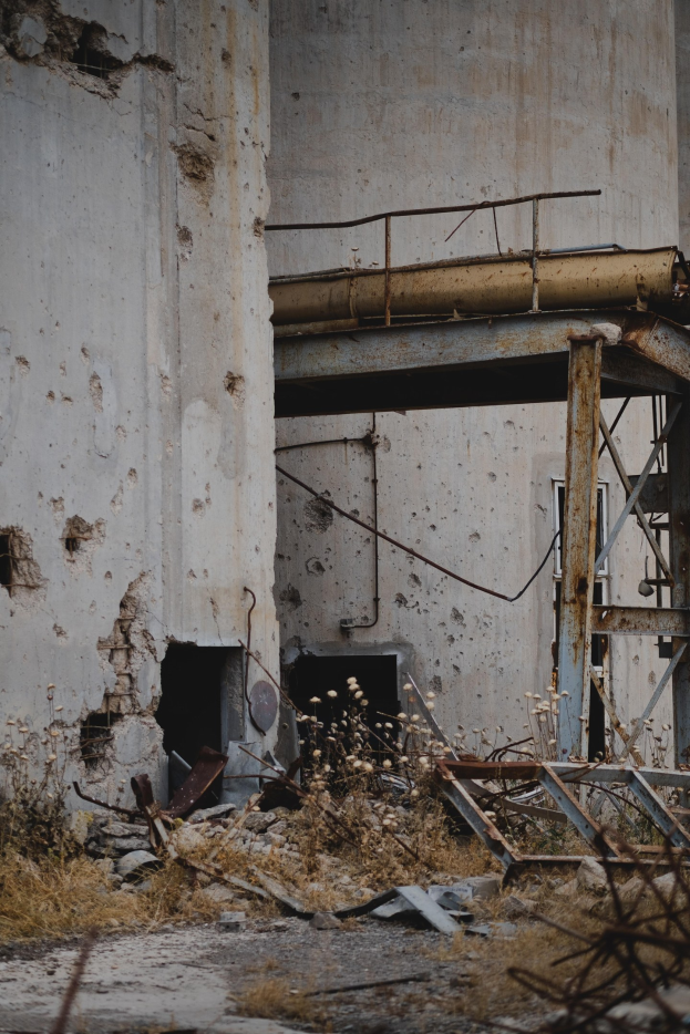 Altes Gebäude mit einer zentralen Metallstruktur, umgeben von Pflanzen, Eisenstäben und Schutt, mit bröckelnden Wänden und zerbrochenen Fenstern.
