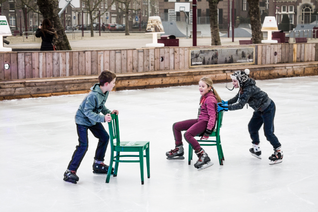 Kinder beim Skifahren vor einem Spielplatz mit drei Kindern und zwei Stühlen in der Mitte und Gebäuden, Bäumen, Bänken, Pfosten und einem Basketballfeld im Hintergrund.