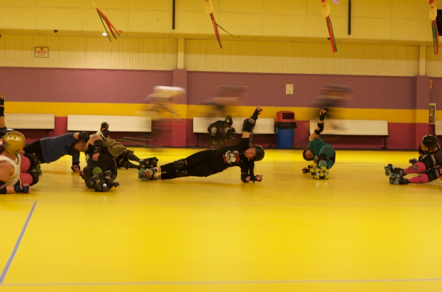 Eine Gruppe von Menschen in Helmen, Ellenbogenschützern, Knieprotektoren und Skateschuhen mit einer Wand und einem Müllcontainer im Hintergrund.
