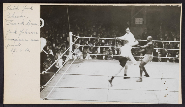 Schwarz-weißes Foto von zwei Boxern, Jack Johnson und Frank Morgan, in einem Boxring mit Zuschauern im Hintergrund.