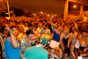 Menschen tanzen im Vordergrund auf einem Konzert mit Lichtern, Pfählen, Bäumen und anderen Gegenständen im Hintergrund.