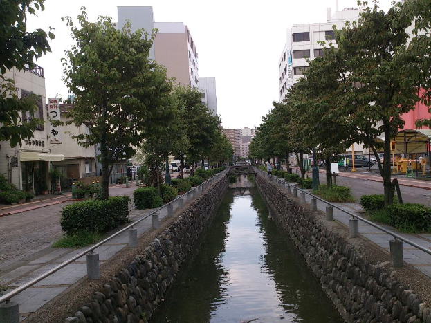 Ein Wasserkanal verläuft durch die Mitte des Bildes, gesäumt von Hecken und Bäumen auf einem Weg, mit Straßen und Gebäuden auf beiden Seiten, fahrenden Autos im Hintergrund und dem Himmel darüber.