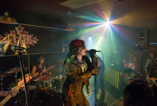 Eine Frau in einer schwarzen Jacke singt in ein Mikrofon auf der Bühne, mit einem Mann in Sonnenbrille und einem Kopfband, der ein Instrument spielt, und einem Plakat mit "LA Guns" im Hintergrund, unter Diskoleuchten und hängenden Lichtern.