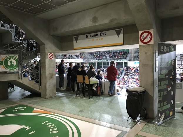 Eine Gruppe von Menschen steht vor einem sitzenden Publikum in einem Stadion, mit Schildern, die "Boston Celtics vs New York Jets" anzeigen, einem Müllcontainer, einer Tafel mit Text, einer Treppe mit Geländern und einem Dach mit Deckenleuchten.