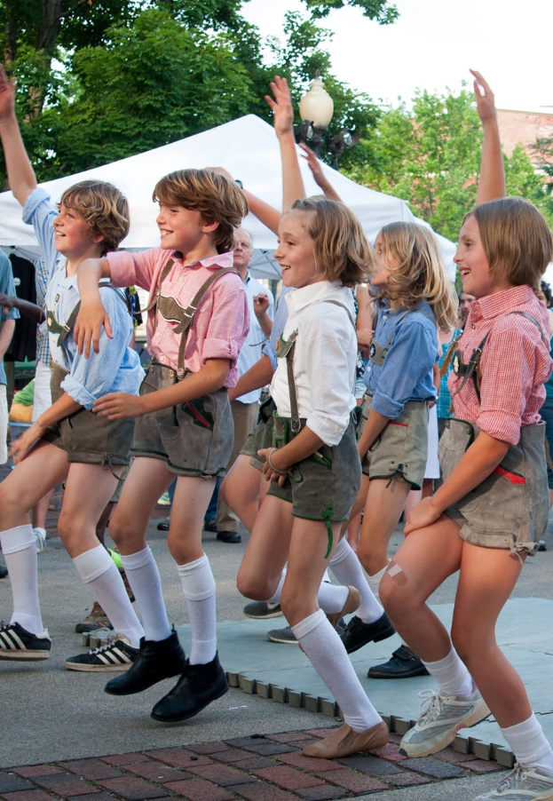 Kinder in traditioneller bayrischer Tracht tanzen auf einer Straße, umgeben von einer Menge, Zelten, Laternenmasten, Bäumen, Gebäuden und einem klaren blauen Himmel.