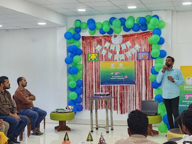 Eine Gruppe von Menschen sitzt auf Stühlen und schaut auf eine Bühne mit Ballons, einem Banner, einer Uhr und anderen Dingen, während jemand auf der Bühne ein Mikrofon hält.