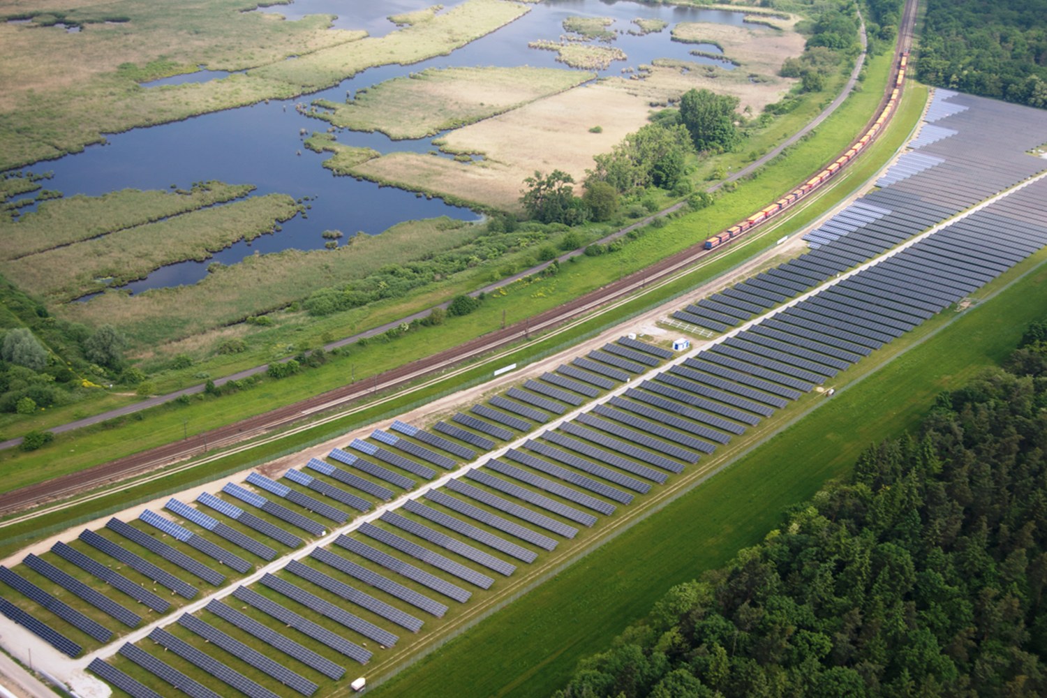Luftaufnahme einer Solarpark mit Solarpanelen, umgeben von Bäumen, Gras, Pflanzen und Wasser, mit einem Zug auf einem Bahngleis sichtbar.