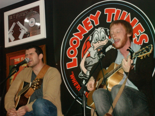 Zwei Personen mit Gitarren, ein Mikrofon davor und ein "Looney Tunes"-Plakat im Hintergrund.