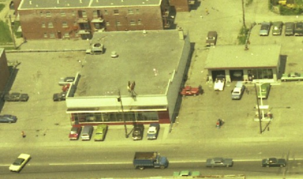 Luftaufnahme einer Stadt mit zahlreichen parkenden Autos um Gebäude herum und einigen Autos auf den Straßen.
