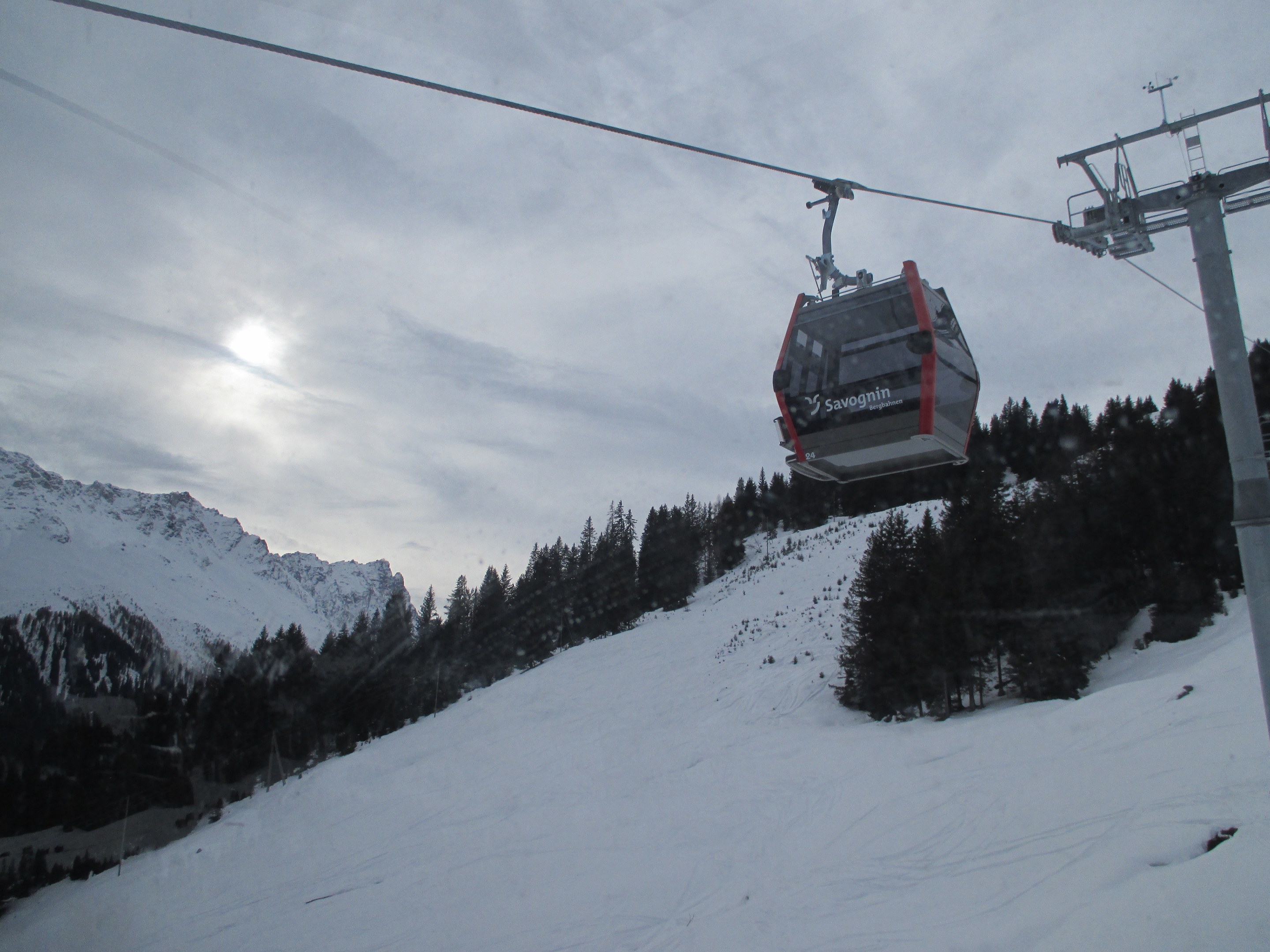 Eine Seilbahn fährt einen schneebedeckten Berg hinauf, mit Bäumen im Hintergrund, unter einem bewölkten, aber sonnendurchfluteten Himmel.