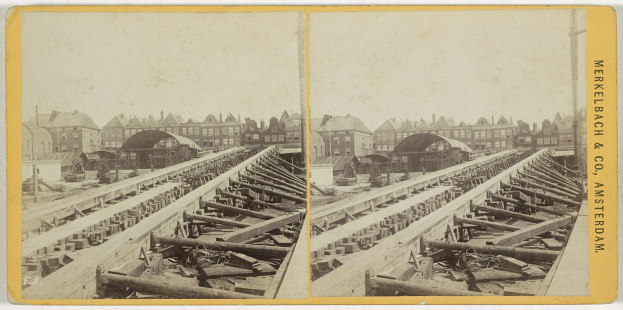 Altes Schwarz-Weiß-Foto einer Schienenstrecke mit Gebäuden im Hintergrund, Text auf der rechten Seite lesbar "Merkelbach & Co, Co. Amsterdam" und verstreute hölzerne Gegenstände im Vordergrund.
