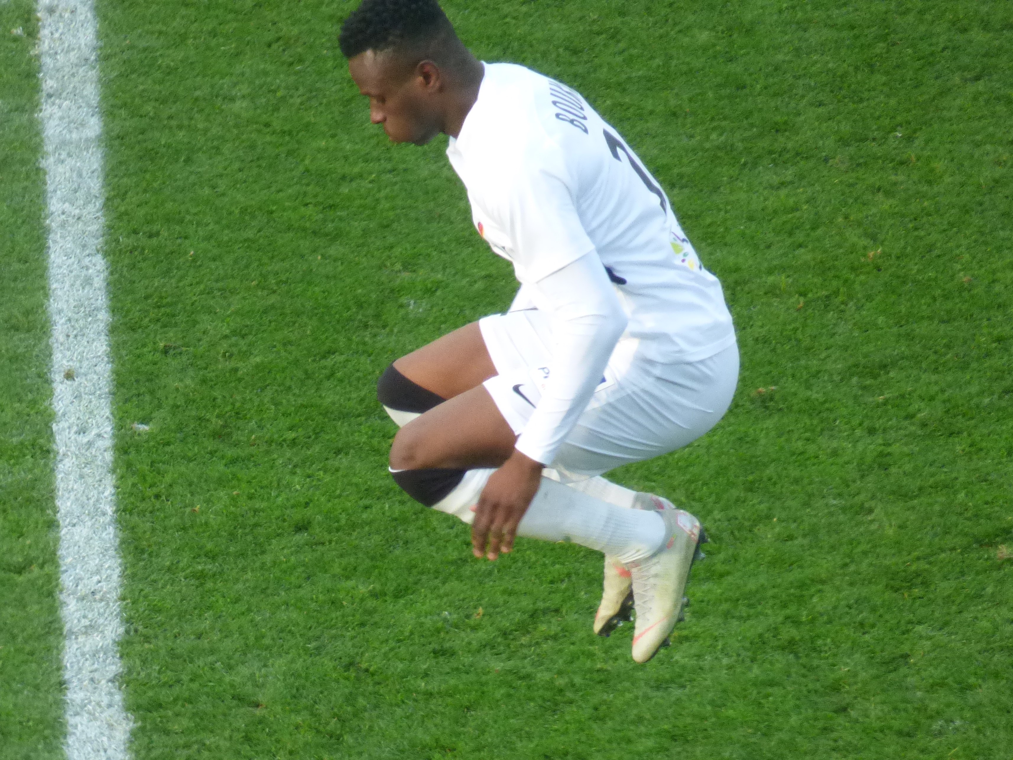 Ein Mann in weißer Uniform kniet auf einem Fußballfeld, trägt Fußballschuhe und eine weiße Linie ist auf der linken Seite des Bildes zu sehen.