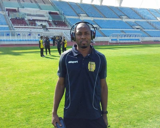 Ein Mann mit Headset, wahrscheinlich Trainer der ghanaischen Fütballnationalmannschaft, steht auf einem Fussballfeld in der Nähe eines Stadions und hält ein Objekt, whährend im Hintergrund Menschen zu sehen sind und der Himmel sichtbar ist.