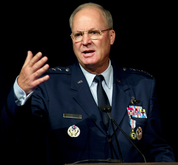 Ein Mann in militärischer Uniform hält eine Rede hinter einem Podium mit zwei Mikrofonen, trägt ein Sakko, eine Krawatte und eine Brille vor einem dunklen Hintergrund.