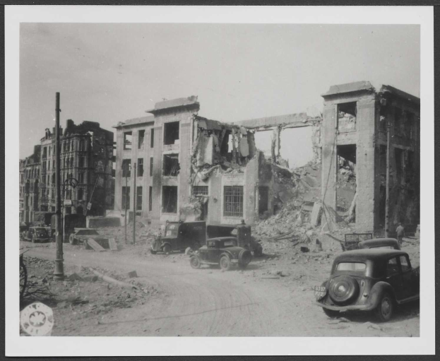 Schwarz-weißes Foto einer Berliner Stadtstraße mit parkenden Autos vor einem stark beschädigten Gebäude, wahrscheinlich von einer Bombenexplosion.