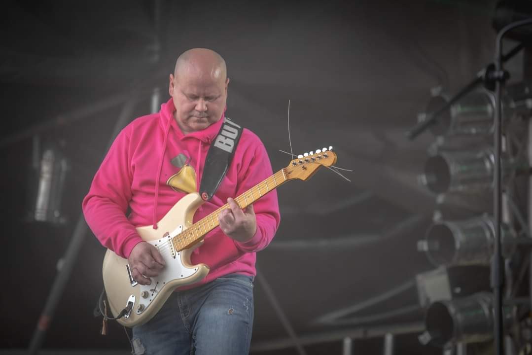Ein Mann in einem pinken Hoodie spielt eine Gitarre auf der Bühne eines Musikfestivals, umgeben von Metallstangen und anderen Gegenständen.