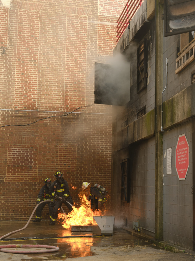 Feuerwehrleute in Helmen nutzen Schläuche, um ein brennendes Gebäude zu löschen, während Rauch aufsteigt; benachbarte Strukturen mit Fenstern und einem Schild sind sichtbar.