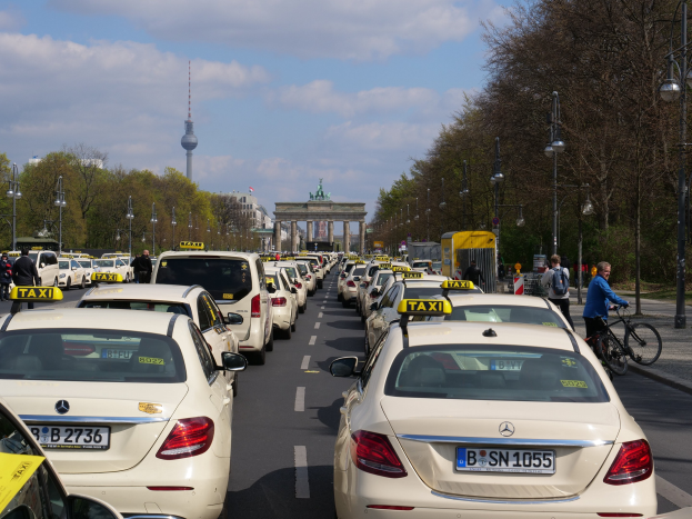 Eine lange Reihe von Taxis, die entlang einer belebten Straße in Berlin, Deutschland, geparkt sind, mit Fahrradfahrern und Fußgängern auf dem Gehweg, flankiert von Bäumen und Laternenpfählen und Gebäuden, einem Bogen und einem Turm im Hintergrund unter einem bewölkten Himmel.